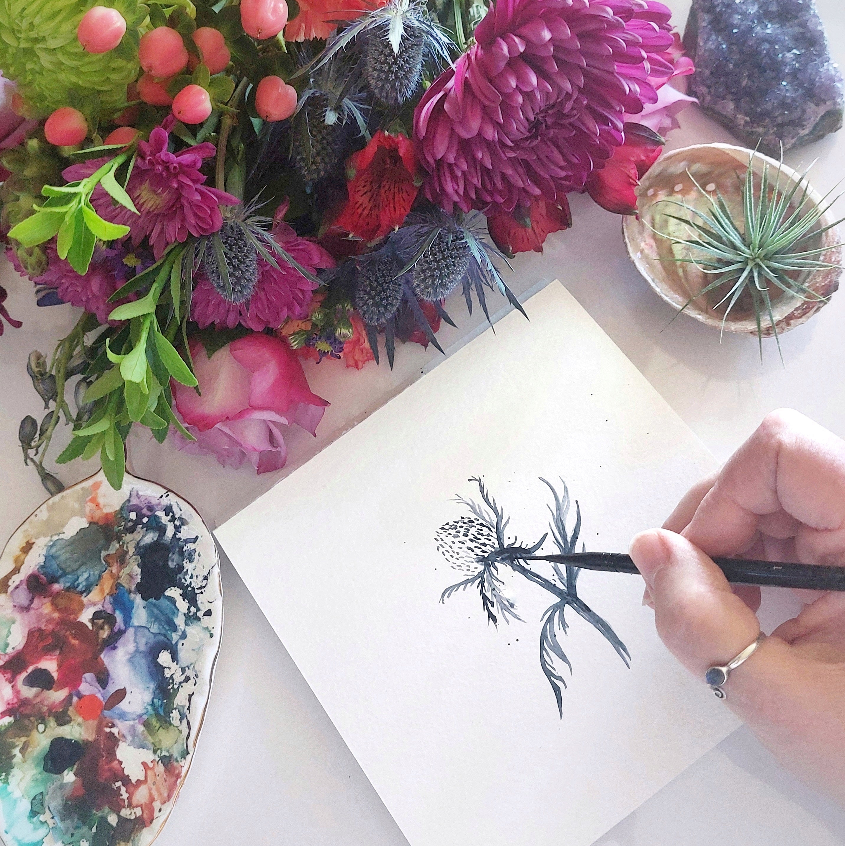 Bridget painting a Scottish Thistle in watercolour surrounded by flowers, an amethyst stone, a seashell and her colourful palette.